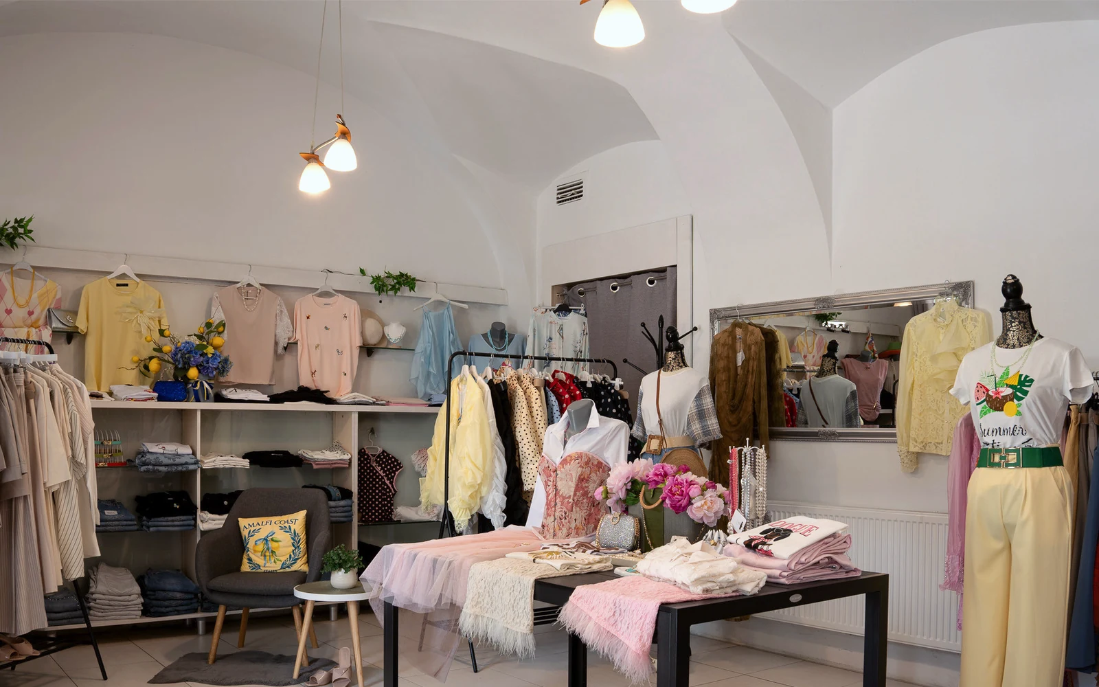 Interiér butiku Amalfi — klenutý strop, stojany s oblečením a stůl s doplňky